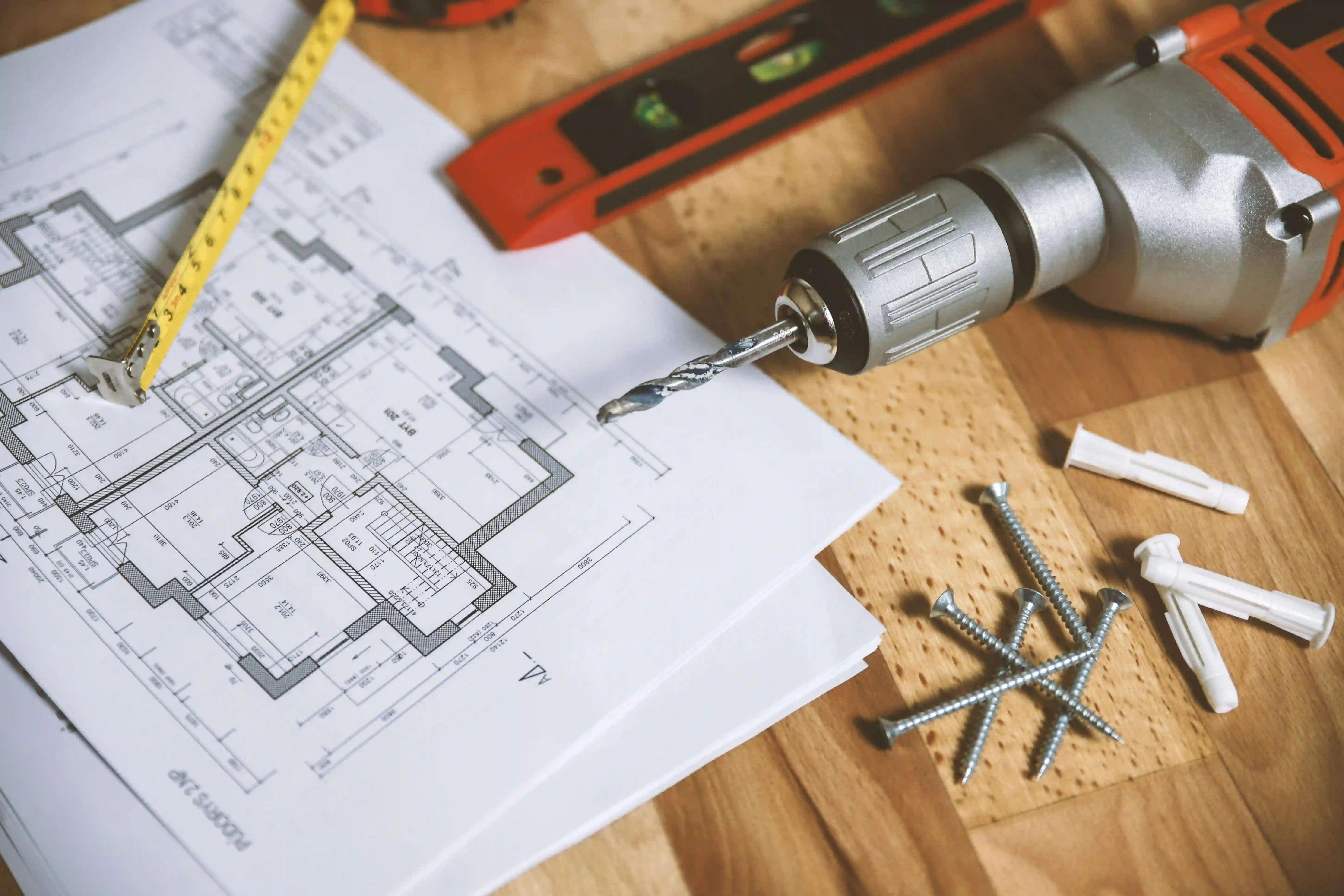 A house blueprint on a wooden table with a tape measure, pencil, and other drafting tools.
